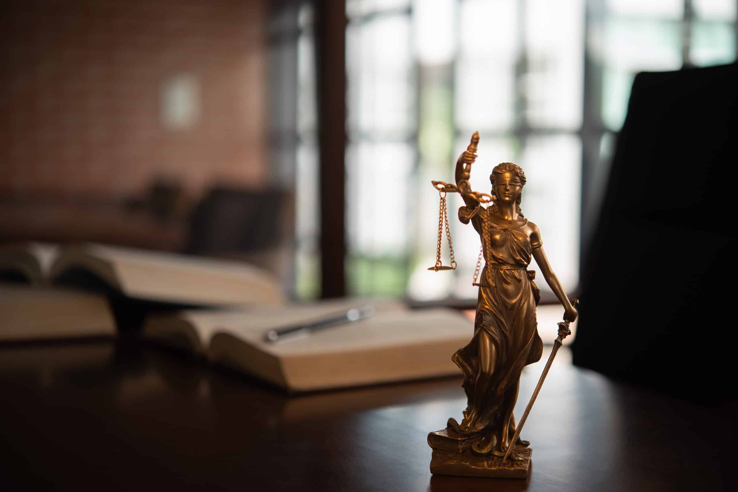 Statuette de la déesse de la Justice (Themis) en bronze, tenant une balance et une épée, sur un bureau sombre près de livres ouverts.